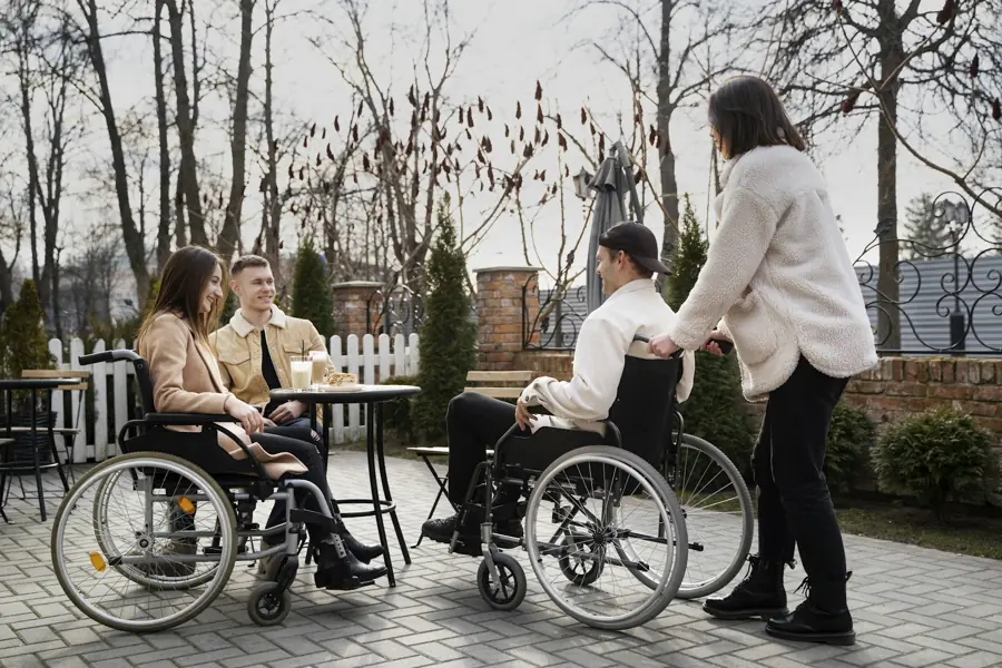 Friends, two of them using wheelchairs, meet at an outdoor cafe table with a support worker.