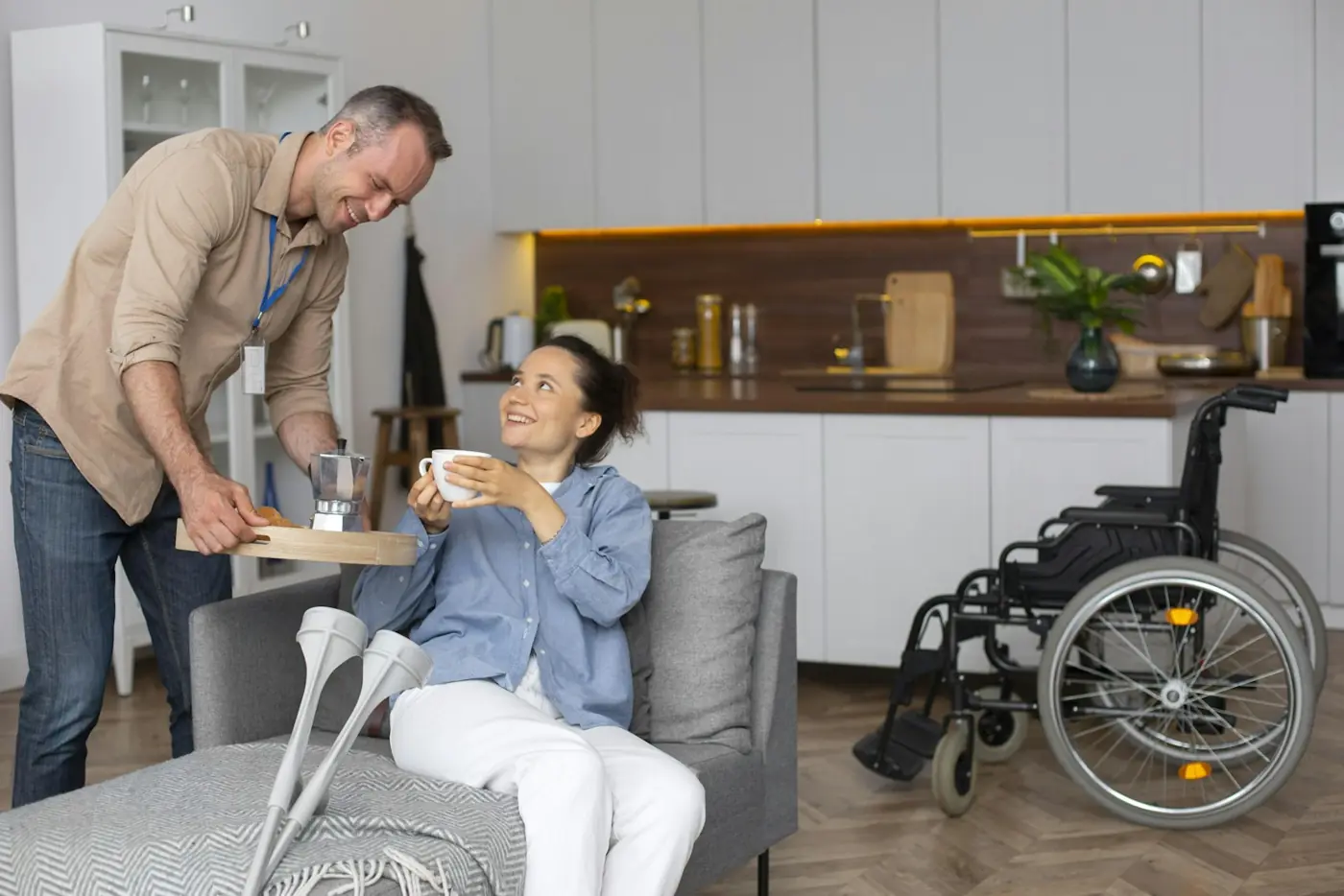 A support worker brings a cup of tea to a smiling woman relaxing on her couch at home.