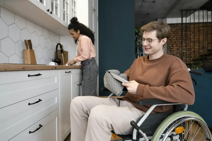 A young man in a wheelchair reads a recipe on a tablet in a bright kitchen.