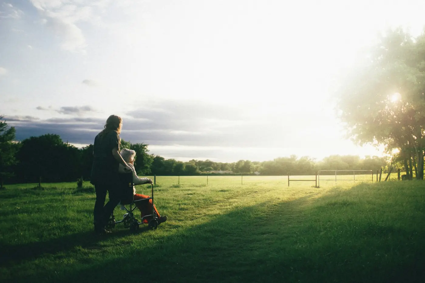 A carer walks beside a person in a wheelchair across a green field at sunset.