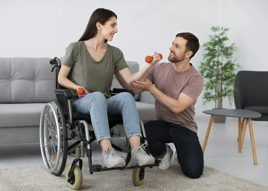 A woman in a wheelchair lifts small weights while a support worker kneels beside her, smiling.