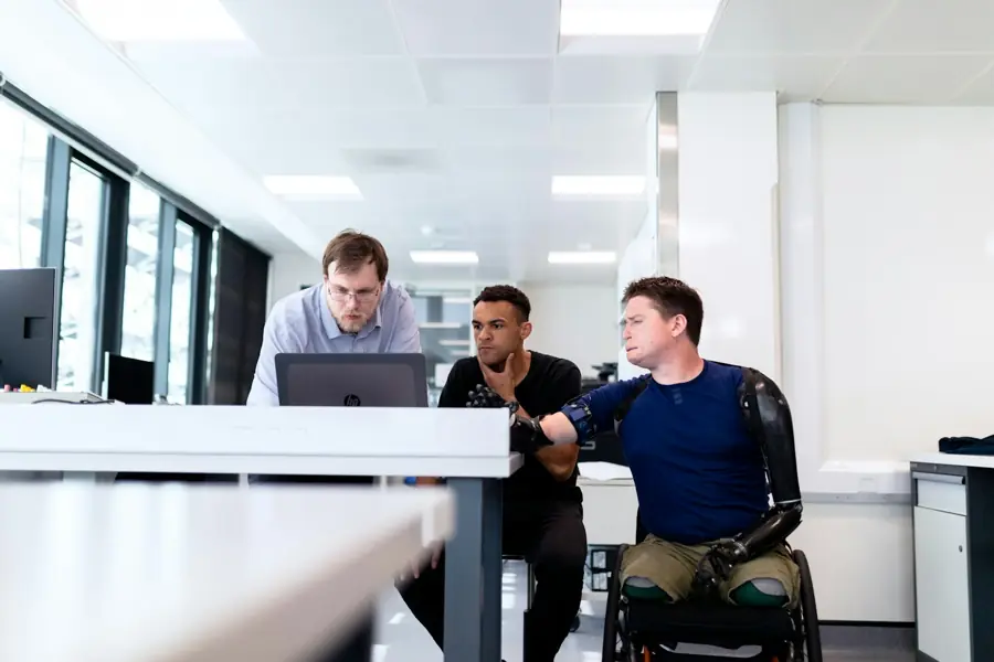A man with prosthetic legs talks with two colleagues at a desk in a modern office.